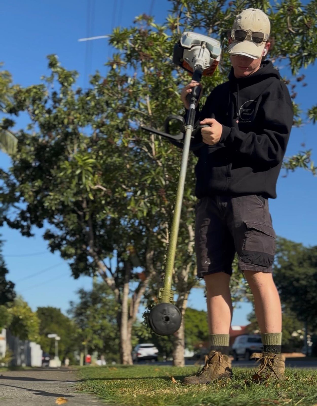 Cooper, owner of Moorooka Cuts, holding a whipper snipper on a suburban footpath